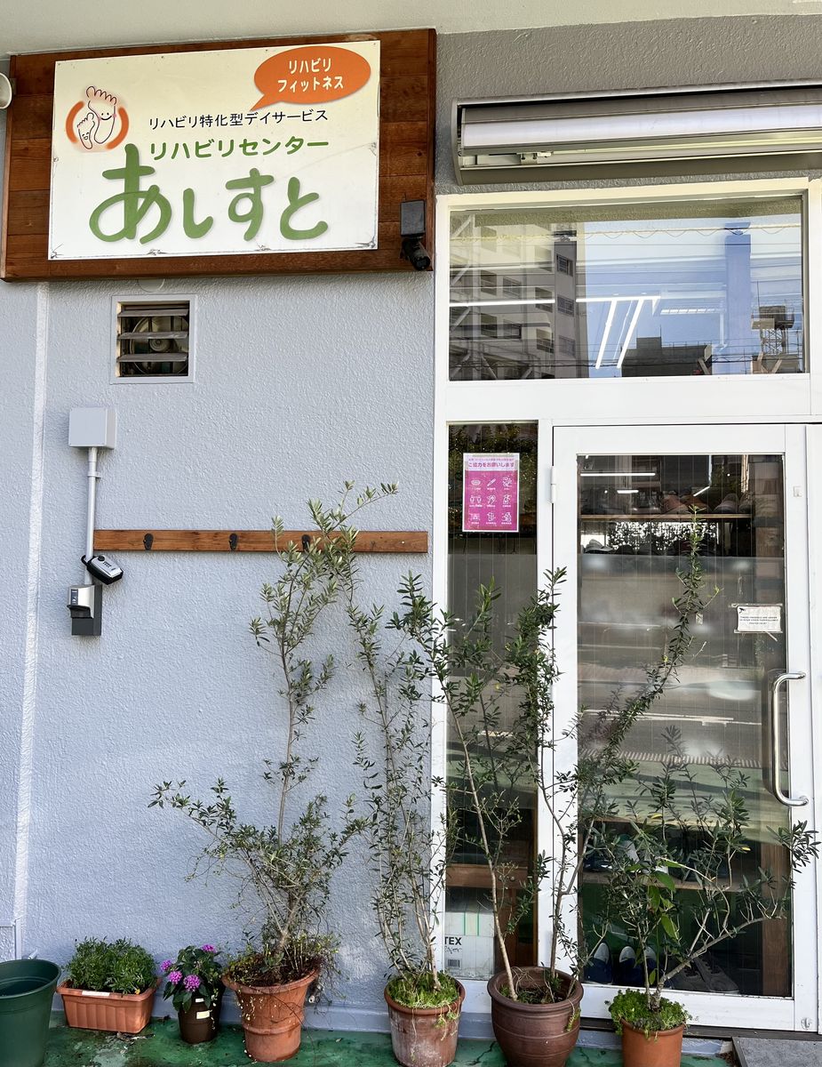「あしすと」の看板がかかった施設の入口と、植木の並ぶ店先